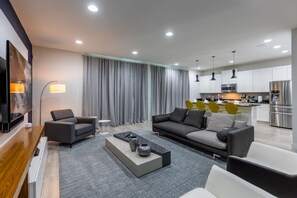 Una sala de estar moderna con sofá, sillones y una mesa de centro. Se observa un área de cocina al fondo con armarios blancos y electrodomésticos de acero inoxidable.