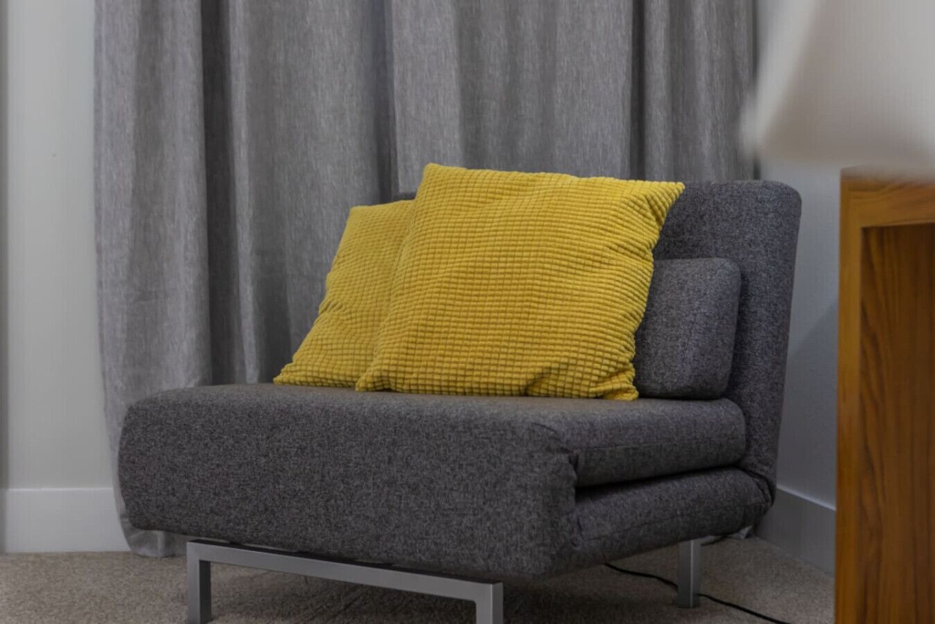 A grey armchair with a mustard yellow cushion against a grey curtain backdrop.