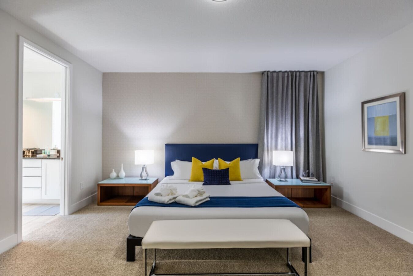 Une chambre d’hôtel avec un grand lit, deux tables de chevet, un banc, une porte donnant accès à une salle de bain et un tableau encadré au mur.