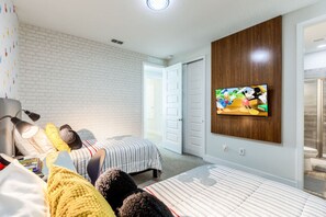 A bedroom with two beds, a TV mounted on the wall, a white brick wall, and a door leading to another room.