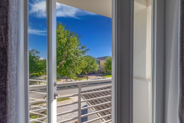 A view from a window with a metal railing, a clear blue sky, and a tree-lined street with buildings in the background.