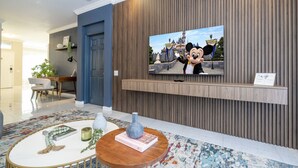 Un salon moderne avec un téléviseur diffusant un dessin animé, un coin repas avec une table et des chaises, et un tapis décoratif.