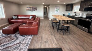 Un salon spacieux avec un canapé en cuir, une table à manger en bois et des tabourets de bar, ainsi qu’un coin cuisine équipé d’appareils électroménagers modernes.