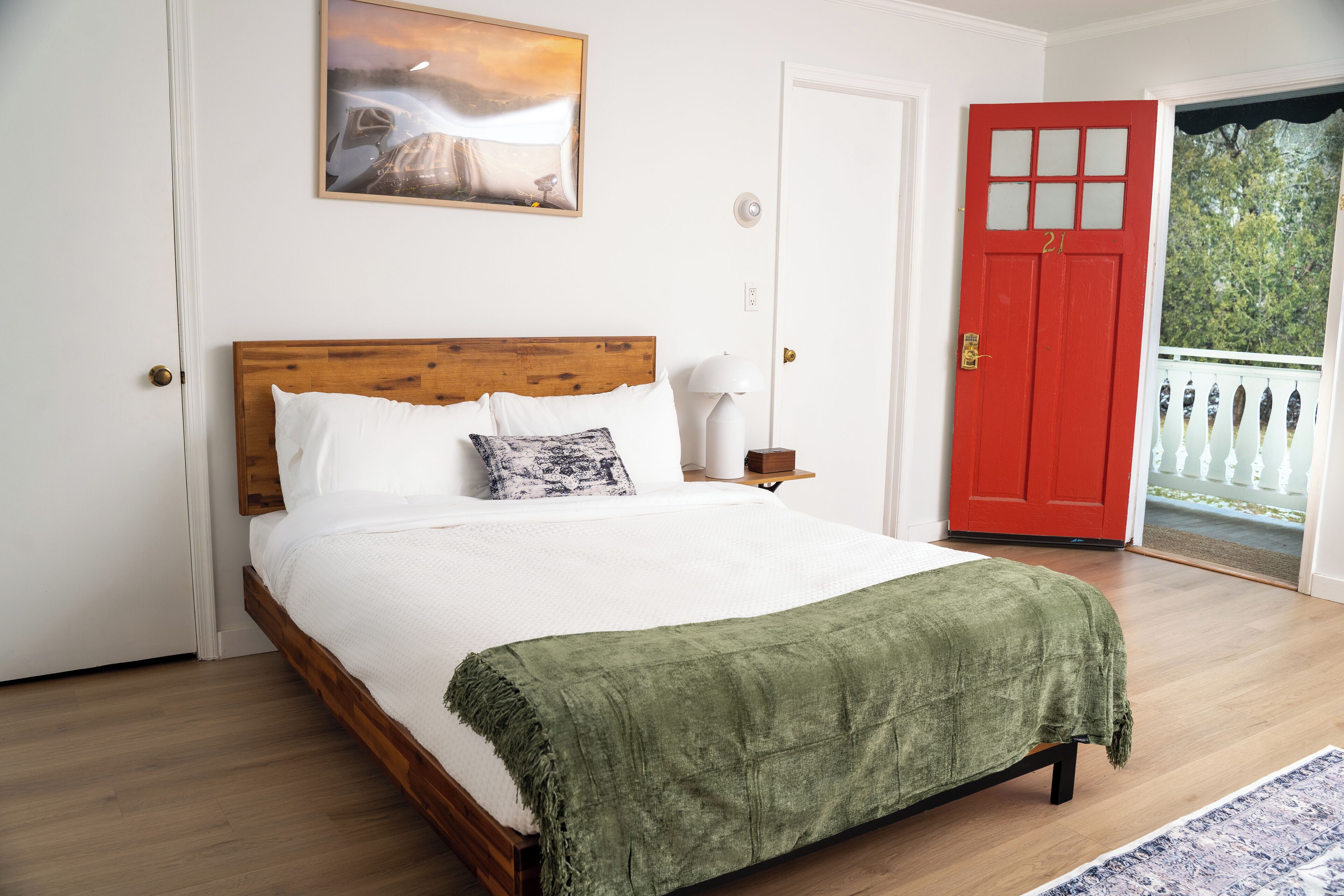 A bedroom with a wooden bed, white bedding, a green throw blanket, a nightstand with a lamp, a red door, and a balcony with white railings.