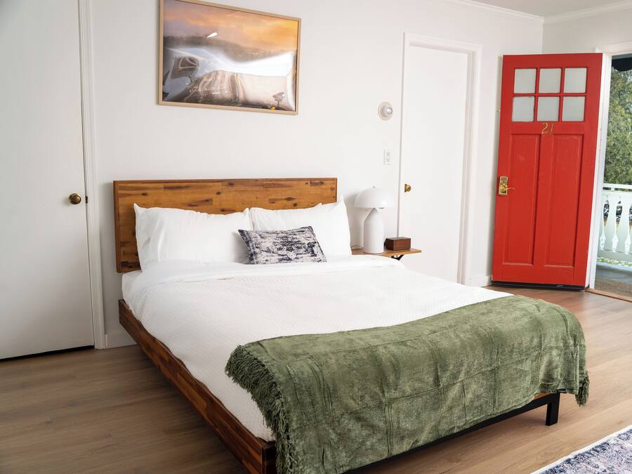 A bedroom with a wooden bed, white bedding, a green throw blanket, a nightstand with a lamp, a red door, and a balcony with white railings.