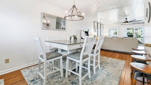 Ein Essbereich mit einem weißen Tisch, Stühlen und einer Chandelier. Im Hintergrund befindet sich eine Couch und ein Deckenventilator.