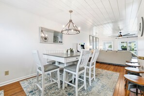 A dining area with a white table, chairs, and a chandelier. There is a sofa and a ceiling fan in the background.