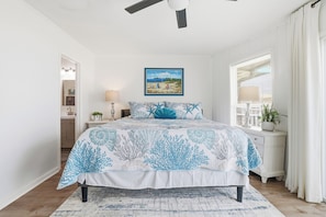 A bedroom with a bed, a ceiling fan, a nightstand, a lamp, a window with curtains, and a painting on the wall.