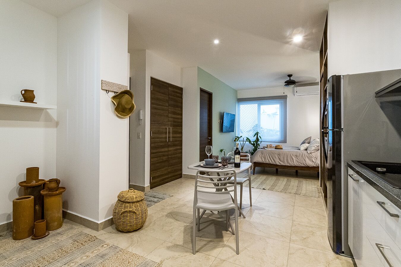 A modern kitchen with a refrigerator, a dining area with a table and chairs, and a bedroom with a bed and window.