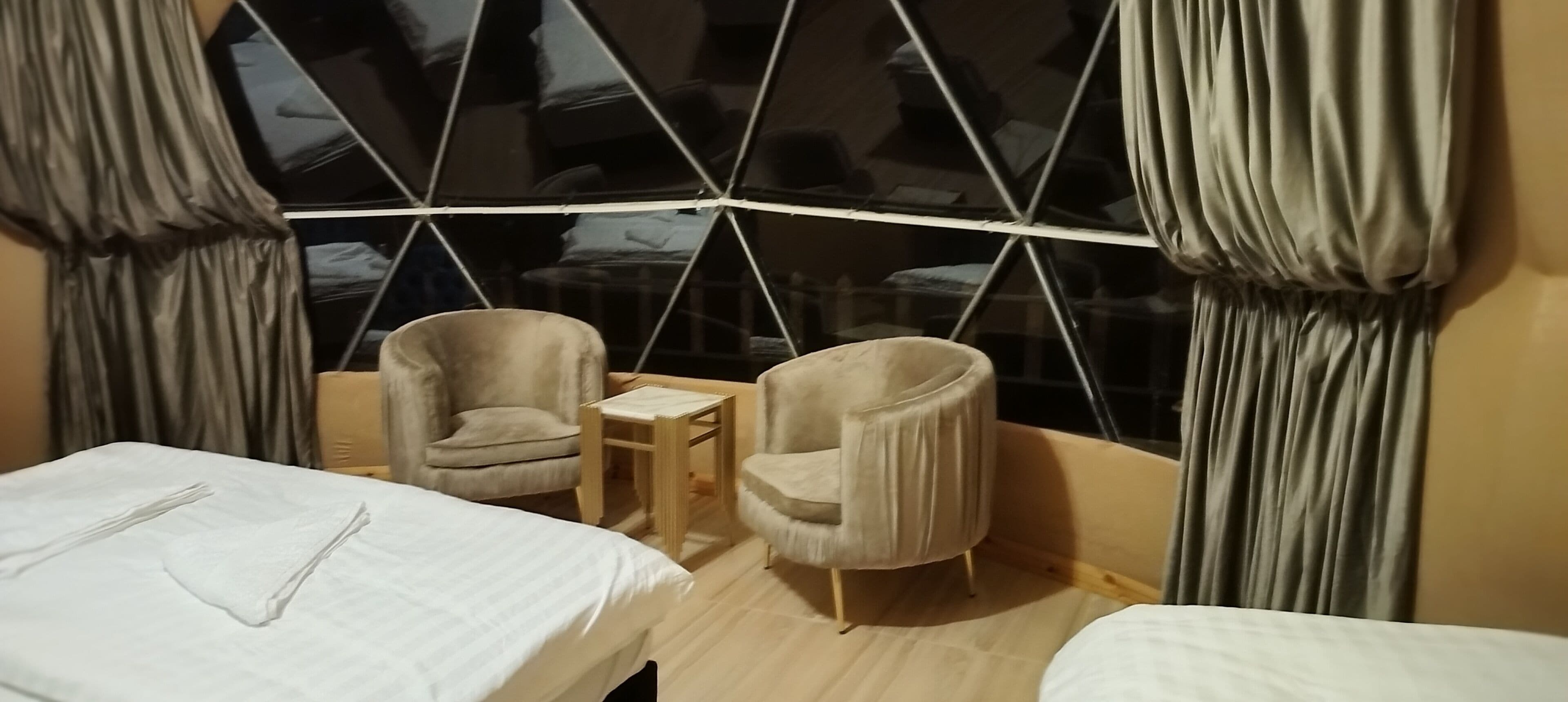 A hotel room with two beds, a small table, two armchairs, and a large window with curtains.