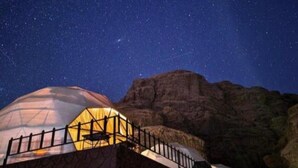 Um observatório abobadado com um passeio cercado, ao fundo um céu estrelado e terreno rochoso.