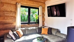 Interior moderno de una cabaña de troncos con un sofá seccional gris, una pequeña mesa redonda y un televisor montado en la pared.
