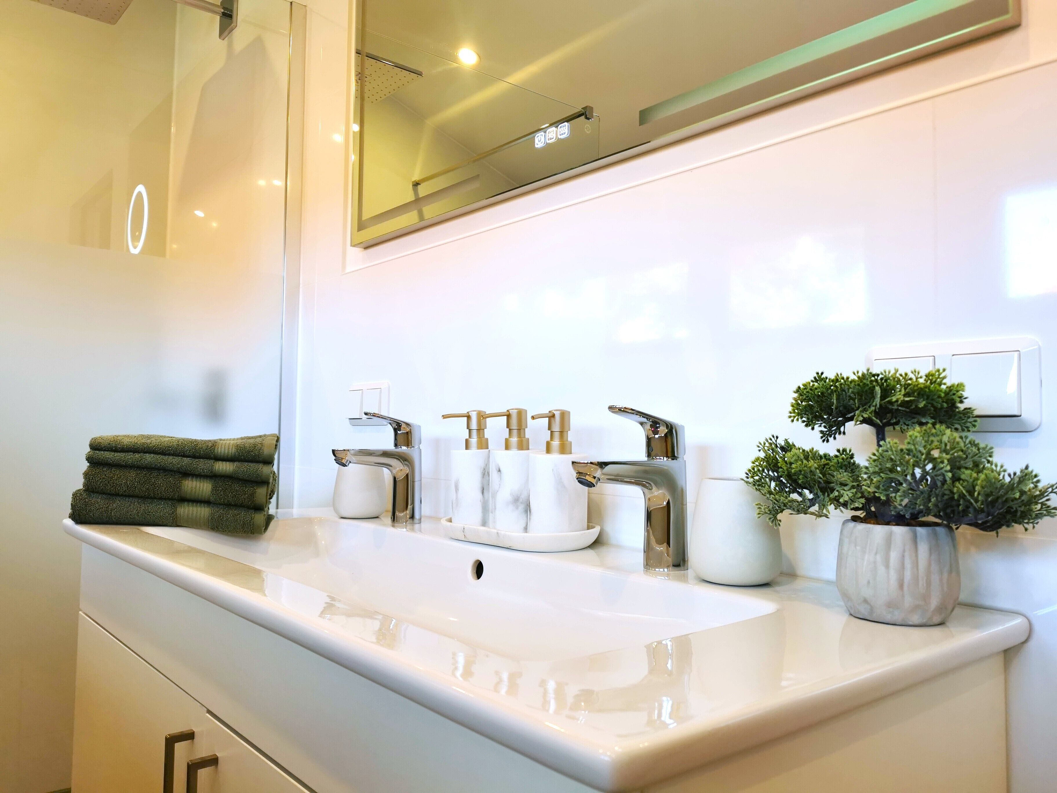 A modern bathroom with a white sink, two gold faucets, and a mirror above. There are towels stacked on the left and a potted plant on the right.