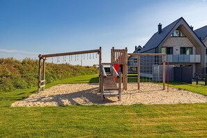 A playground with a slide and climbing structures, surrounded by sand and grass, with a modern house in the background.