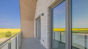 A balcony with a view of a yellow field, a wooden slatted wall, and a glass door.