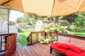 A covered outdoor deck with a wooden railing, a red cushioned bench, and a view of a backyard with a white mobile home and trees. - Family-Friendly Waterloo Abode w/ Deck! (Waterloo)
