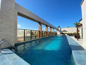 Uma piscina coberta moderna com água azul cristalina, rodeada por um muro de tijolos e painéis de vidro, com vista para edifícios ao fundo.