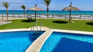 En swimmingpool med utsikt över havet och stranden, omgiven av gräs och palmer.