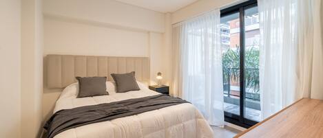 A modern bedroom with a large bed, a bedside table with a lamp, and a view of the outdoors through a glass door.