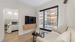 Ein modernes Wohnzimmer mit einer Couch, einem an einer Holzkonsole montierten Fernseher und einem großen Fenster mit Blick auf Gebäude.