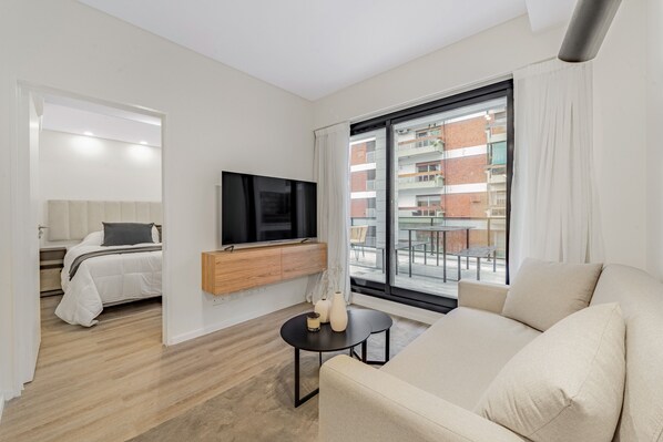 A modern living room with a sofa, a TV mounted on a wooden console, and a large window with a view of buildings.