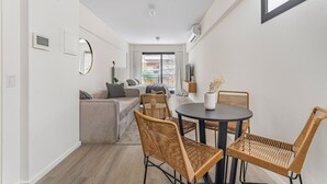Ein modernes Wohnzimmer mit einer Couch, einem Esstisch und Korbstühlen. Ein Fenster mit Vorhängen, ein Heizkörper und ein Blumentopf sind ebenfalls zu sehen.