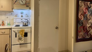 A kitchen with a stove, oven, and a clock above the stove. There is a door to the right and a wall with a quilt hanging. - Clean, cozy, 1-bedroom basement apartment: 4-month rental, 2nd semester. (Toronto)