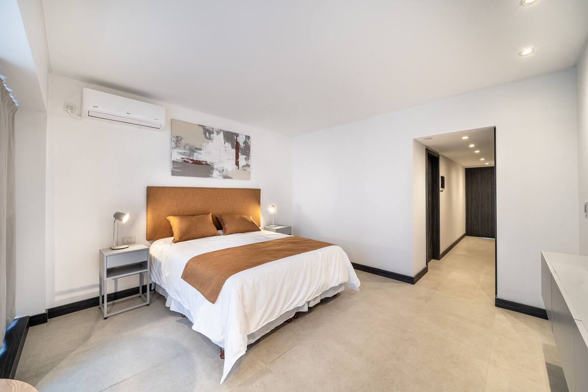 Une chambre d’hôtel moderne dotée d’un grand lit, de tables de chevet, d’un appareil de climatisation et d’un couloir menant à une autre chambre.