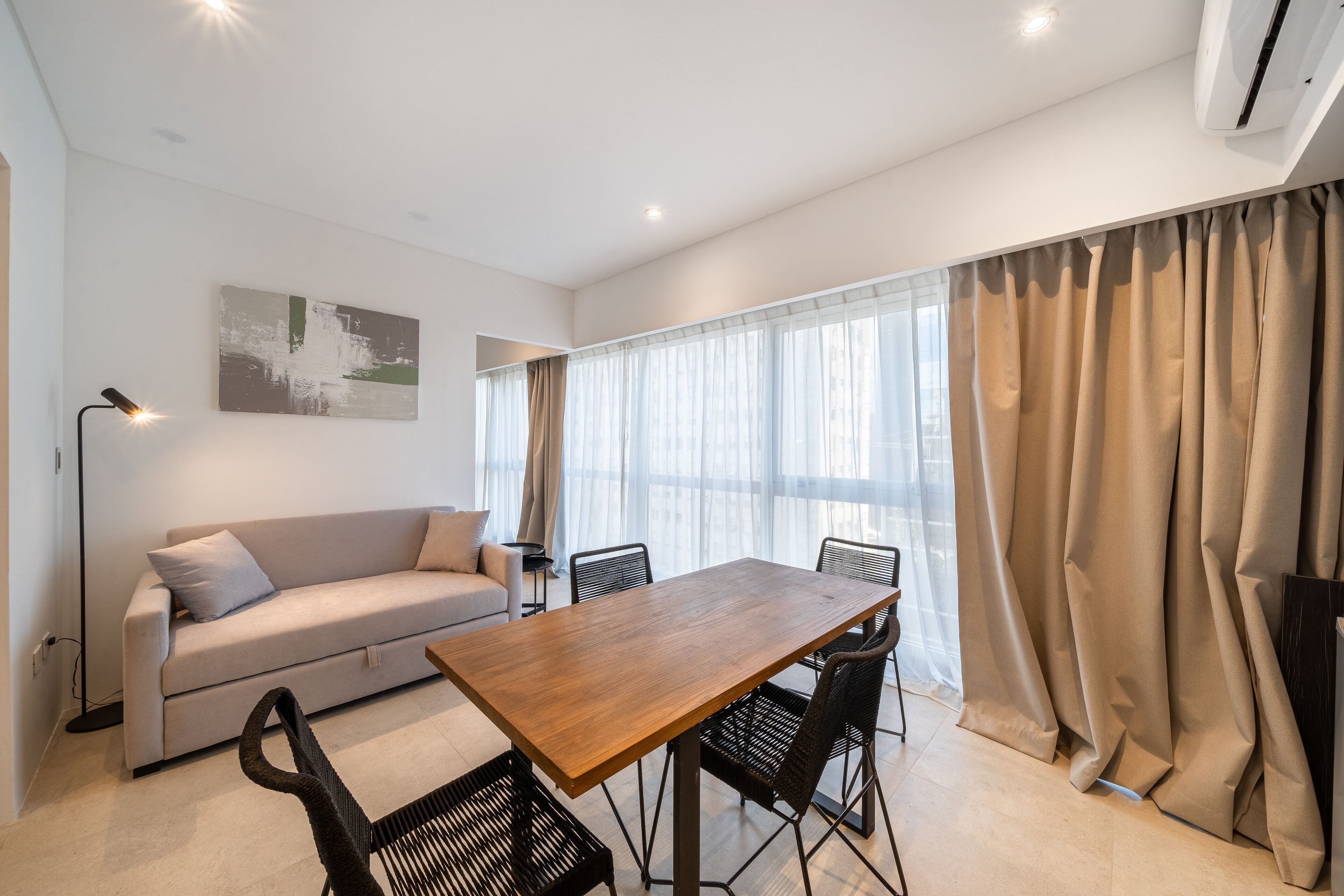 A modern living room with a wooden dining table, black chairs, a beige sofa, and a large window with sheer curtains.