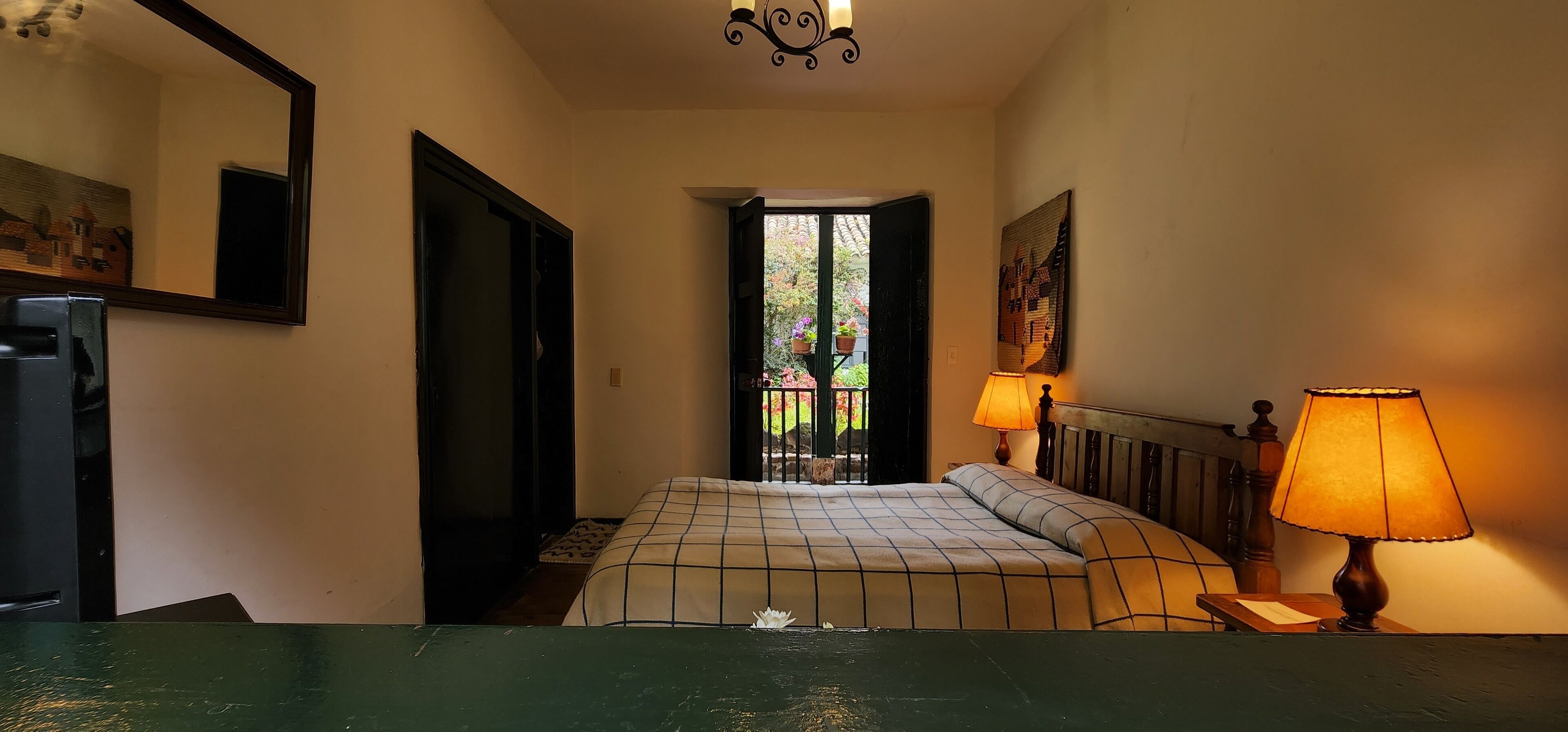 A bedroom with a bed, two bedside lamps, a mirror, and a view of a garden through an open door.