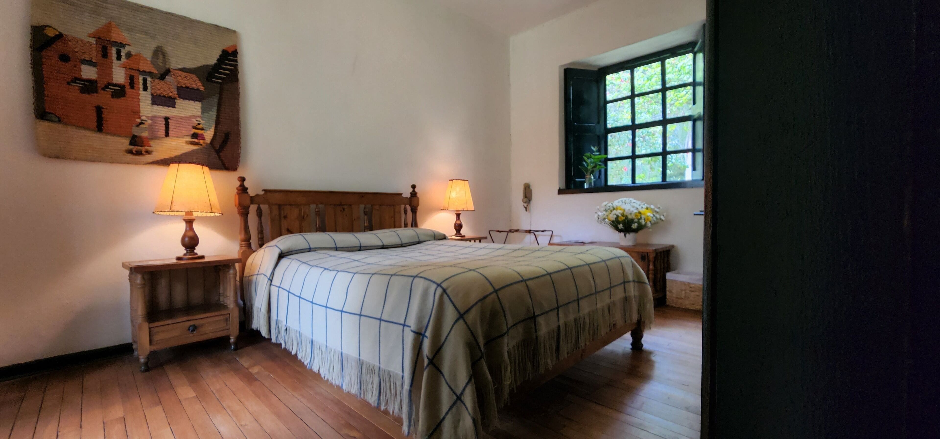 Ein Schlafzimmer mit einem hölzernen Bett, zwei Nachttischen mit Lampen, einem Wandbild, einem Fenster mit Blick und einer Vase mit Blumen.