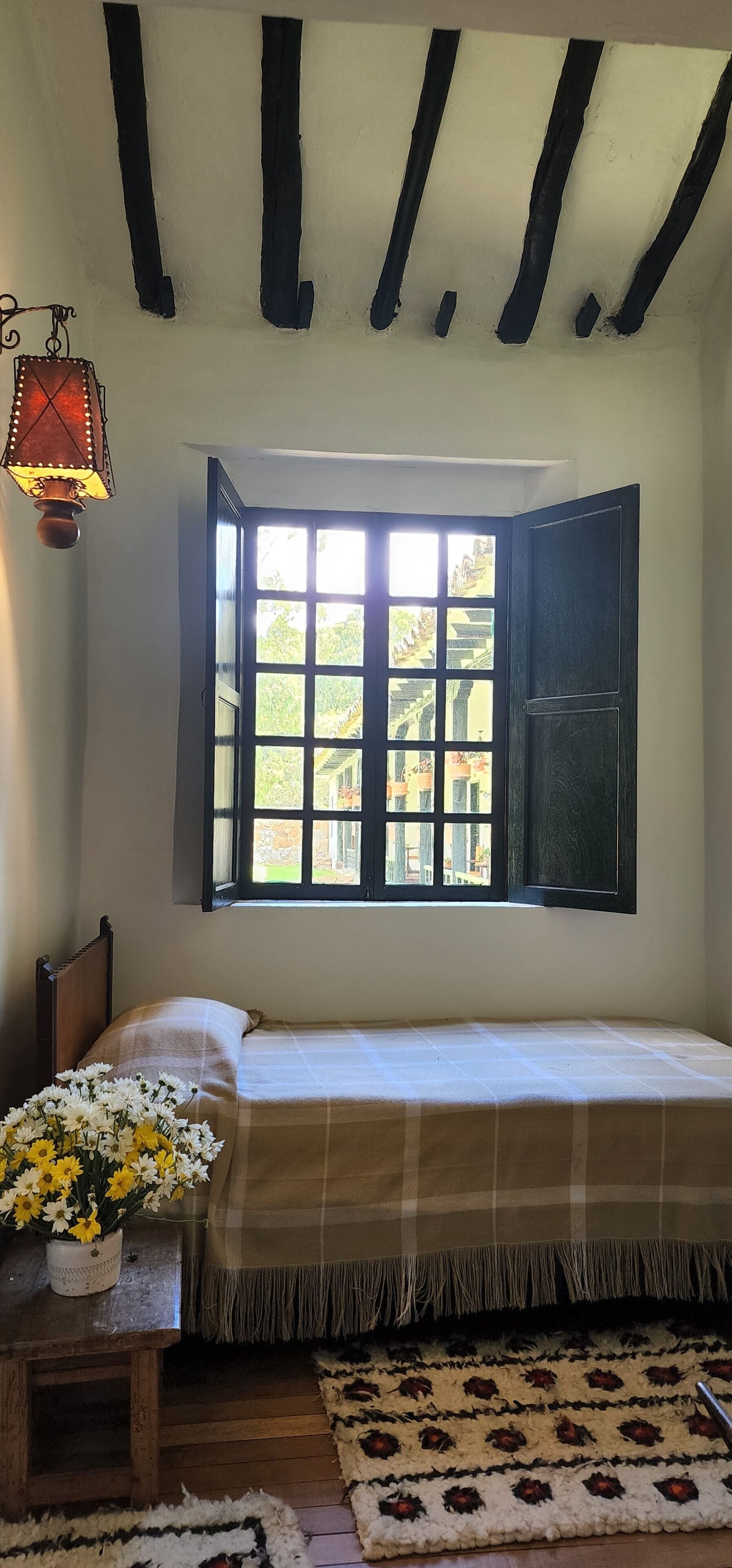 Una habitación con cama, ventana con vistas a la vegetación, una mesita con flores y una lámpara colgante.