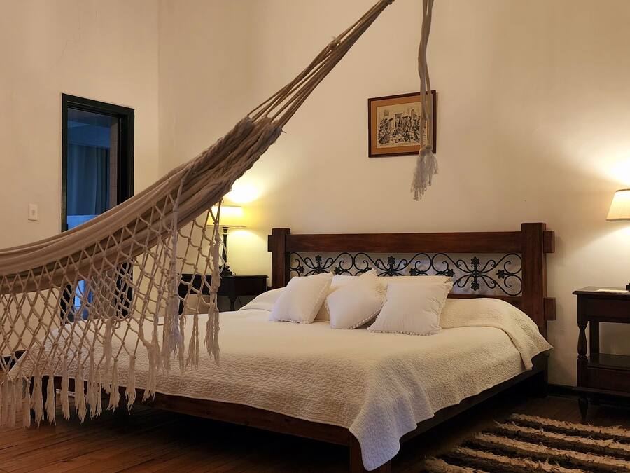 Un dormitorio con una hamaca, una cama con ropa de cama blanca, dos lámparas de noche, una mesita de noche de madera y un cuadro enmarcado en la pared.