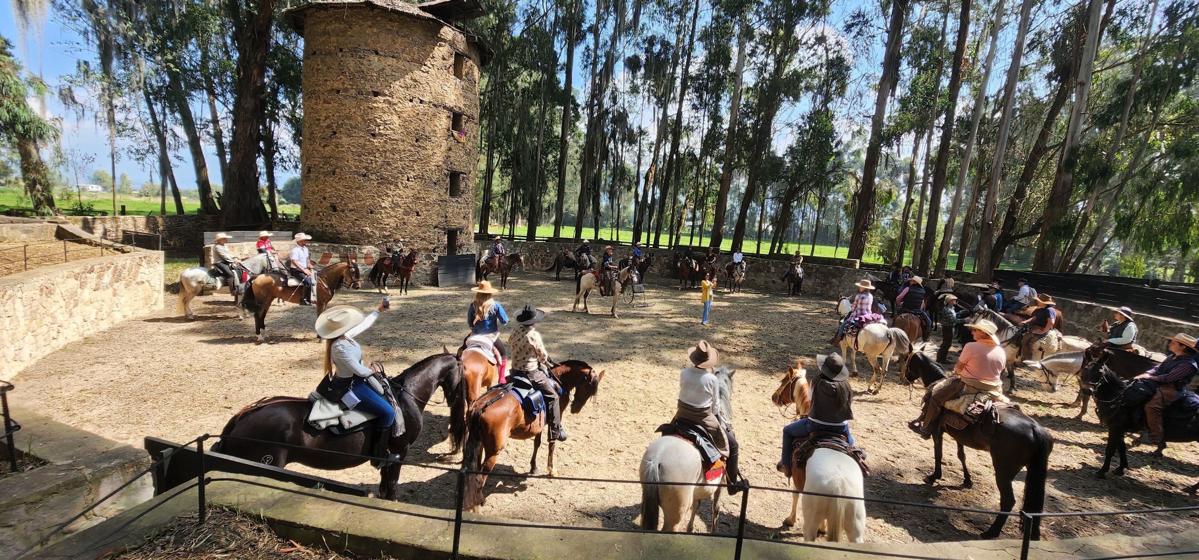 Un grupo de personas a caballo en una arena al aire libre, con una estructura de piedra y árboles al fondo.