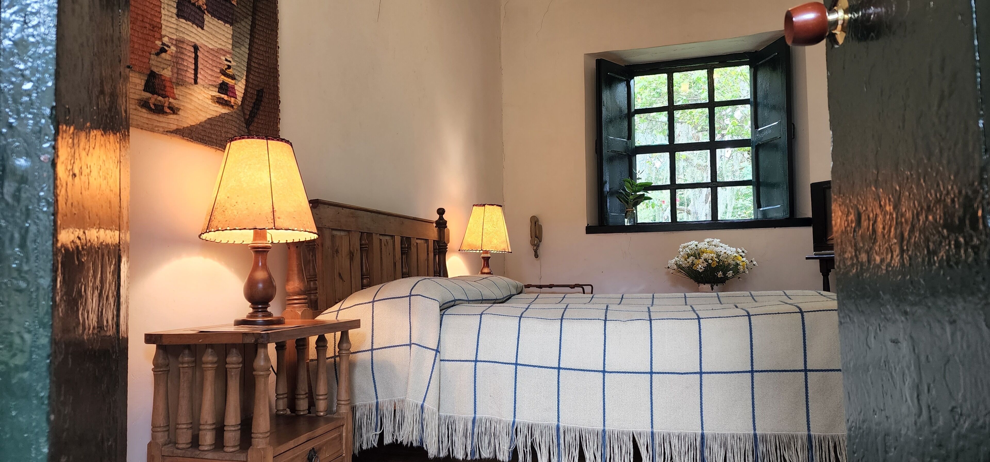 A cozy bedroom with a wooden headboard, a checkered bedspread, two bedside lamps, a window with a view of greenery, and a vase with flowers on the nightstand.