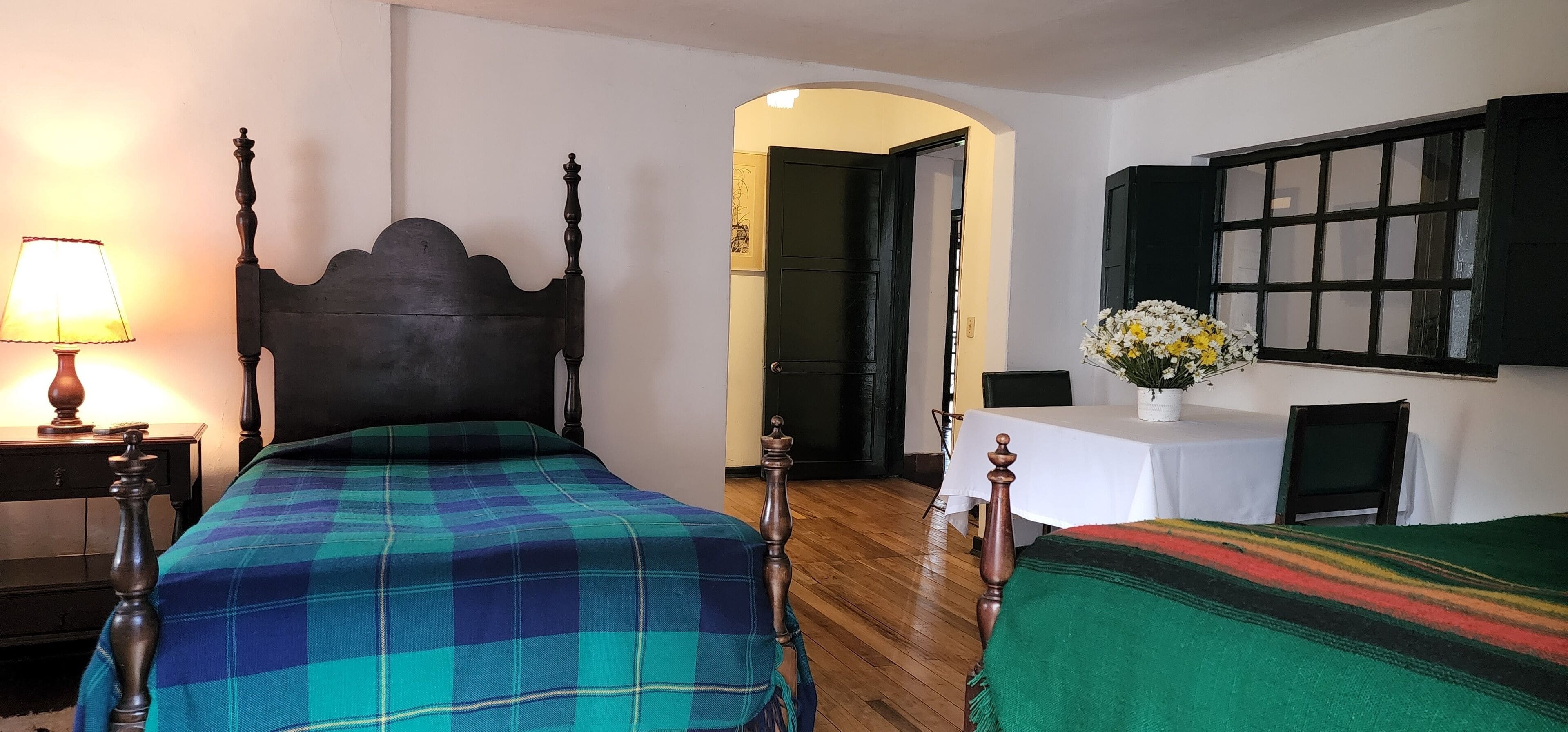 Una habitación con dos camas, una mesa con un florero, una lámpara y una ventana con contraventanas.