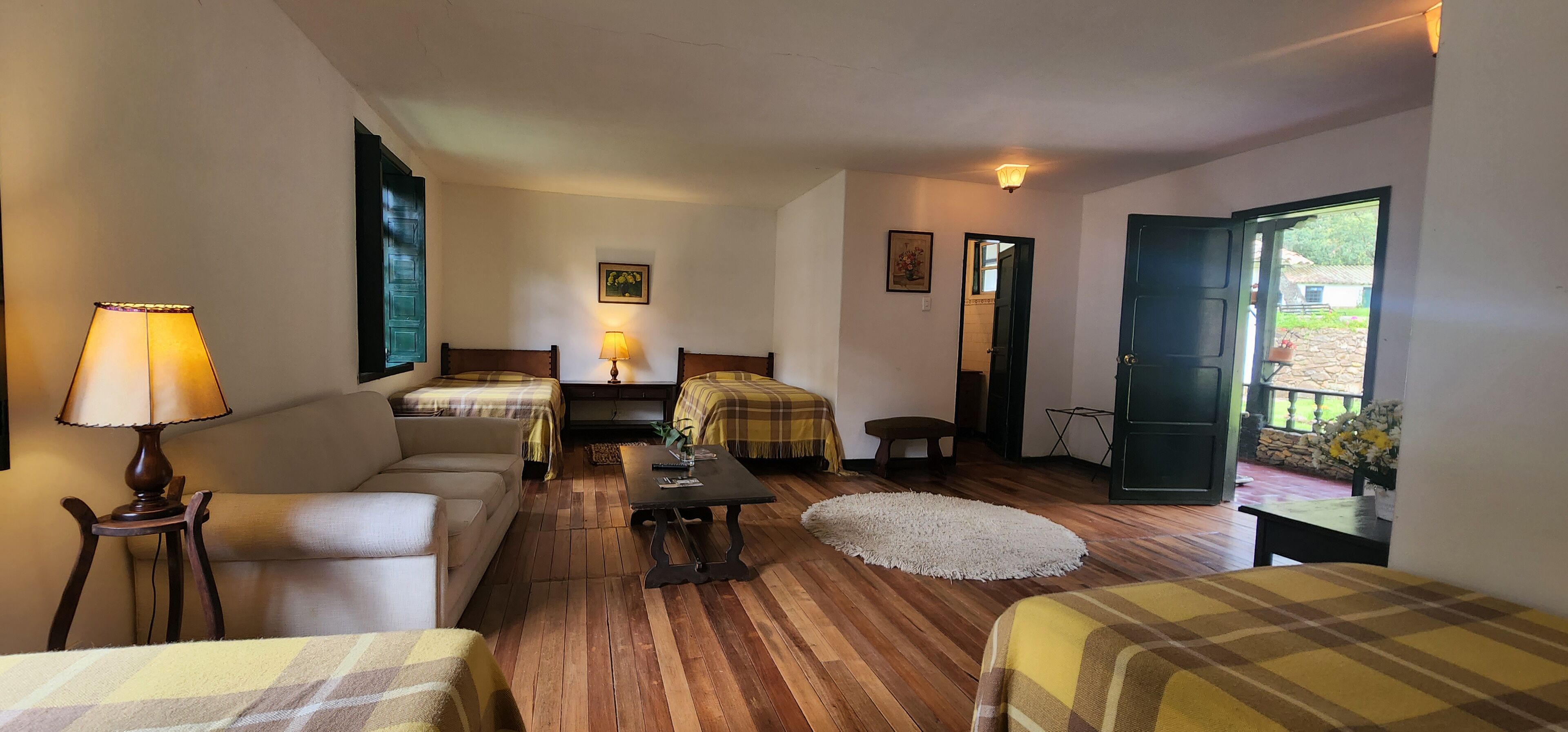 A cozy room with two beds, a sofa, a small table, and a lamp. There is a door leading to an outdoor area with flowers.