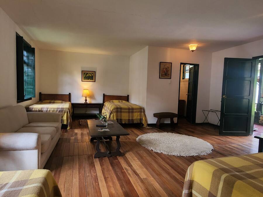A cozy room with two beds, a sofa, a small table, and a lamp. There is a door leading to an outdoor area with flowers.