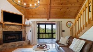 A cozy living room with a stone fireplace, a leather sofa, a flat-screen TV, and a large window offering a view of the outdoors. - The Sanctuary in Blue, Long Range Mountain Views (Blue Ridge)