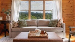 A cozy living room with a beige sofa, a small table with a tray, and a view of a forest through large windows. - The Sanctuary in Blue, Long Range Mountain Views (Blue Ridge)