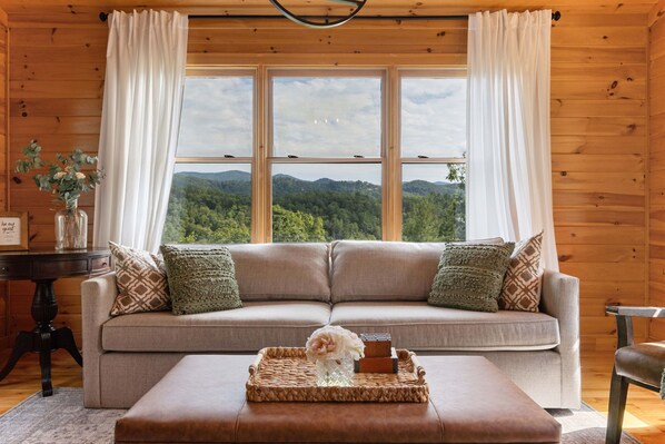 A cozy living room with a beige sofa, a small table with a tray, and a view of a forest through large windows.