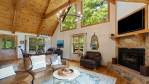 A spacious living room with a stone fireplace, leather sofas, a television mounted on the wall, and large windows offering a view of the outdoors. - The Sanctuary in Blue, Long Range Mountain Views (Blue Ridge)