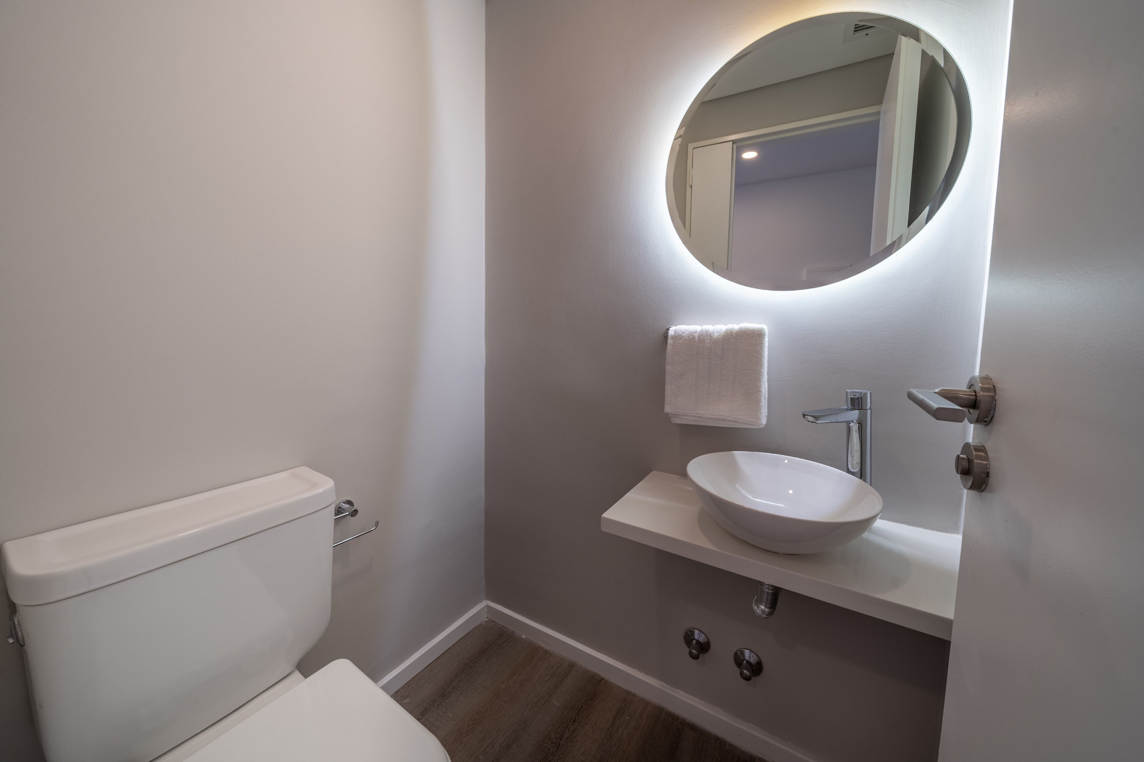 A modern bathroom with a white toilet, a round mirror with a light, and a white sink with a towel hanging on the wall.