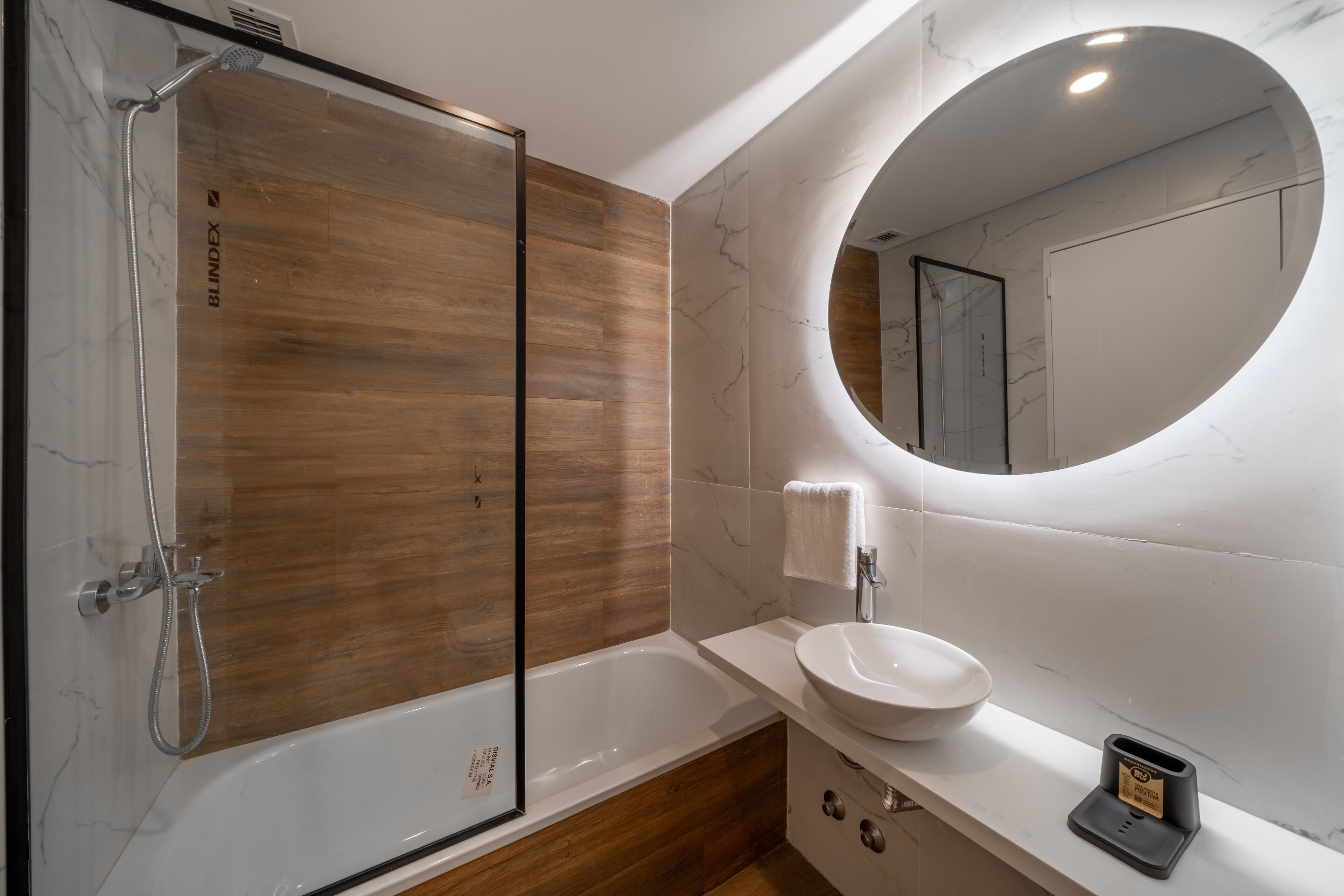 Une salle de bain moderne dotée d’une douche à parois de verre, d’un meuble-lavabo en bois, d’un miroir rond et d’un lavabo encastré.