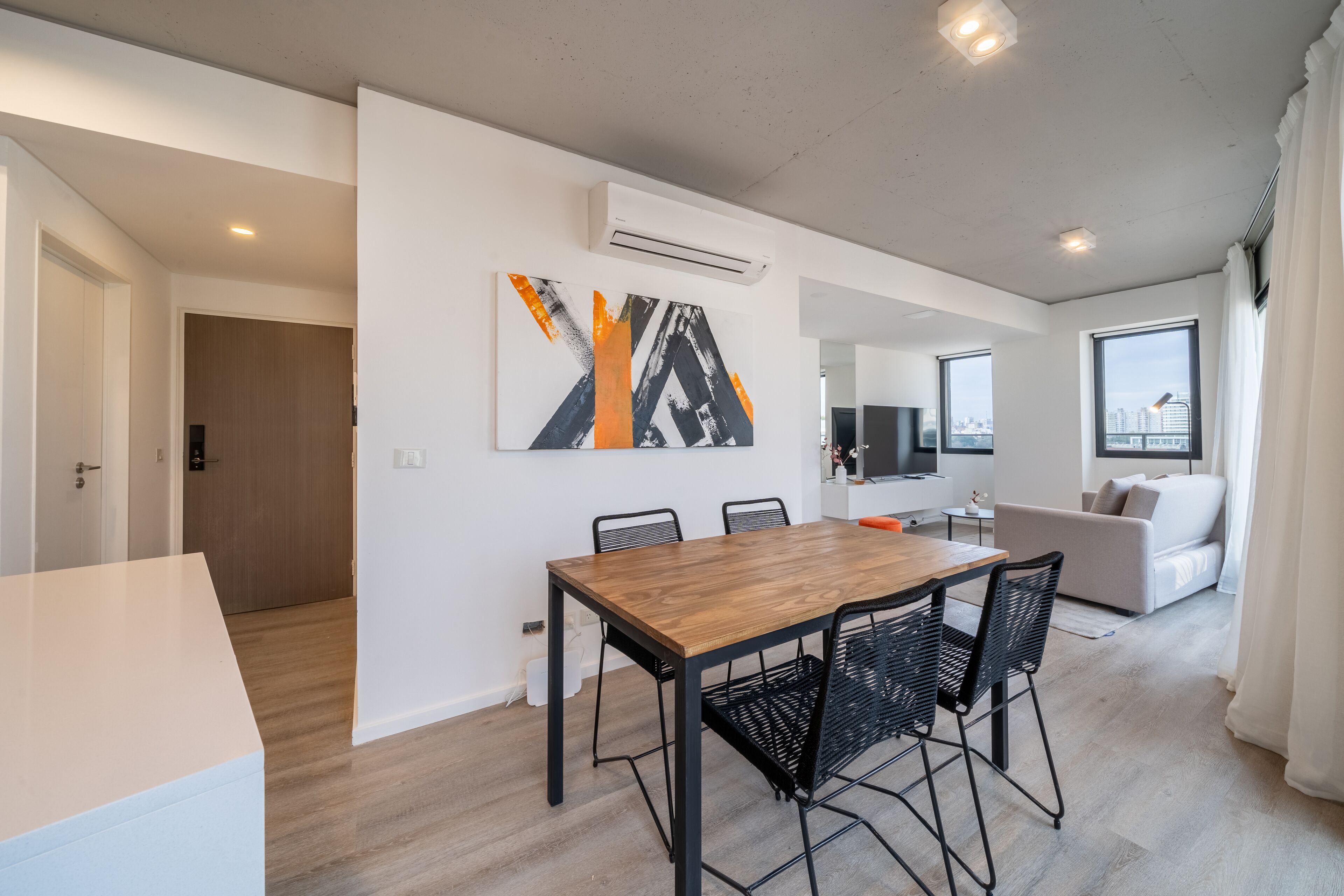 A modern living room with a dining area, featuring a wooden table and black chairs, a sofa, and a large window with a city view.