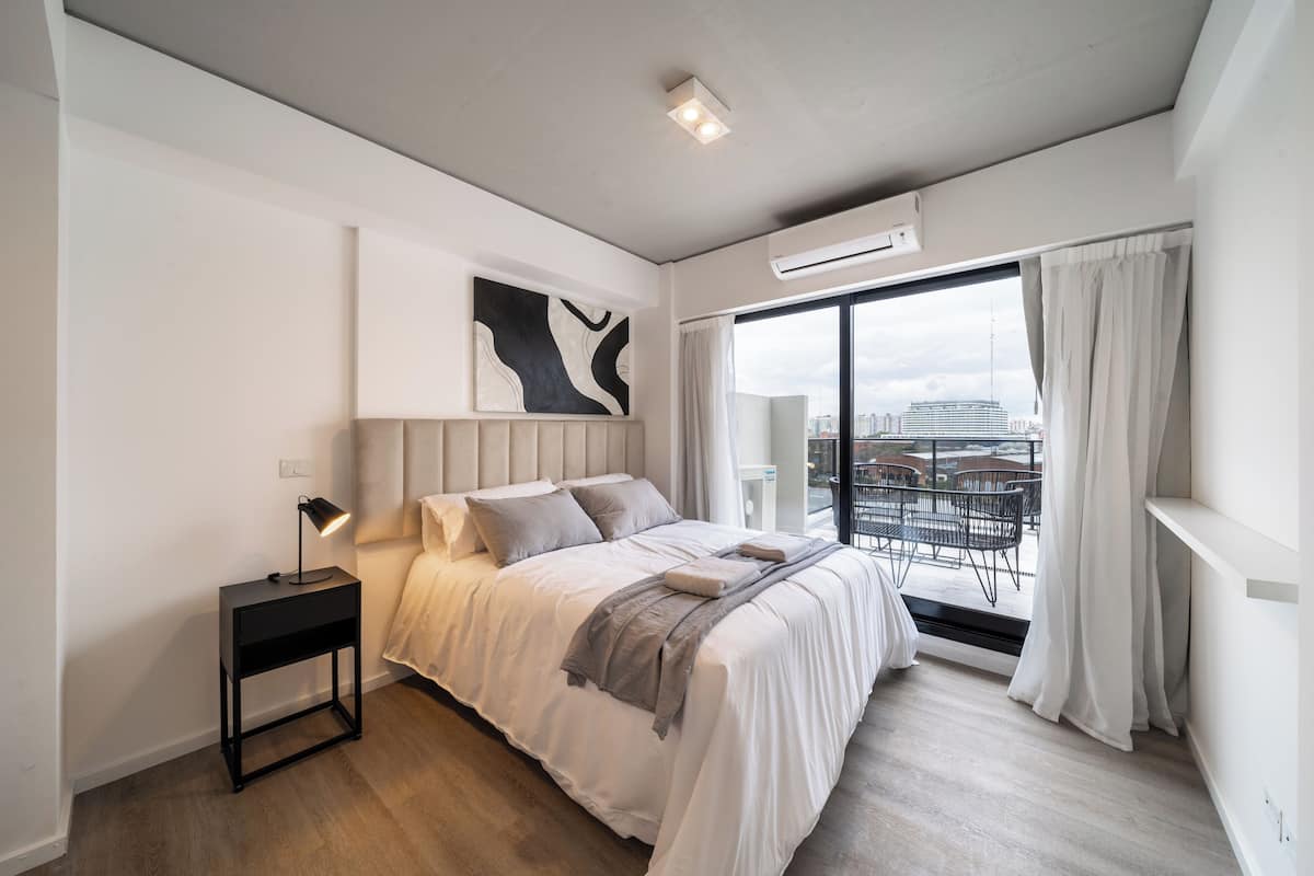 Een moderne slaapkamer met een groot bed, een nachtkastje met een lamp, uitzicht op de stad door een groot raam en een plafondlamp.