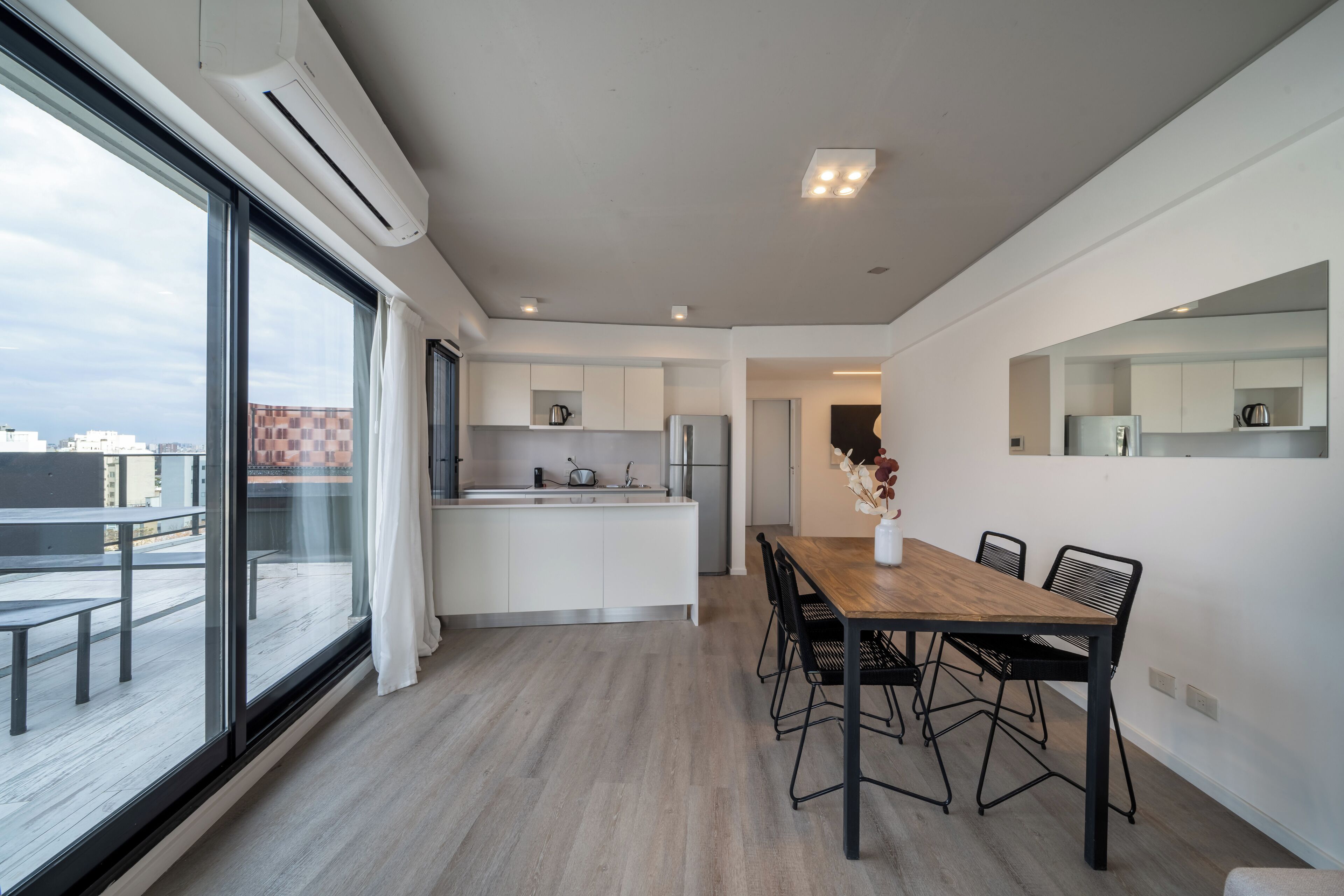 A modern kitchen with white cabinets, a wooden dining table, and black chairs. There is a large window providing a view of the city.