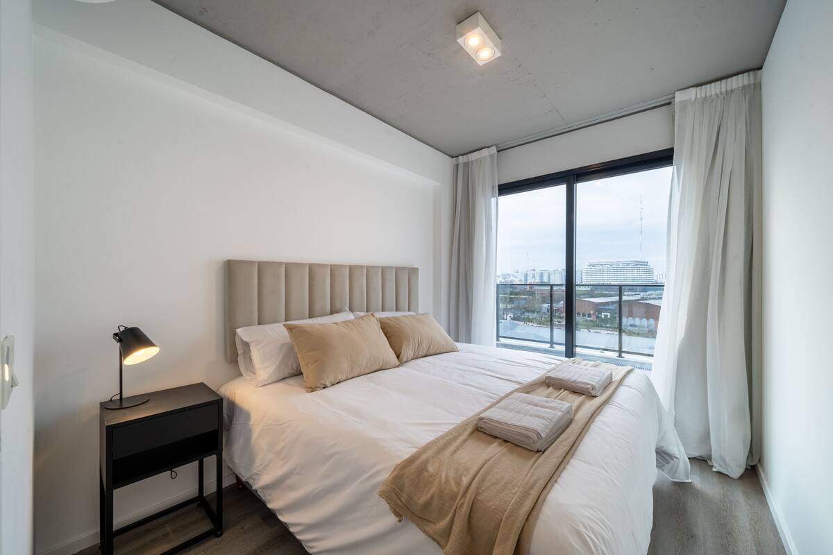 Een moderne hotelkamer met een groot bed, een nachtkastje met lamp en uitzicht op de stad door een groot raam.