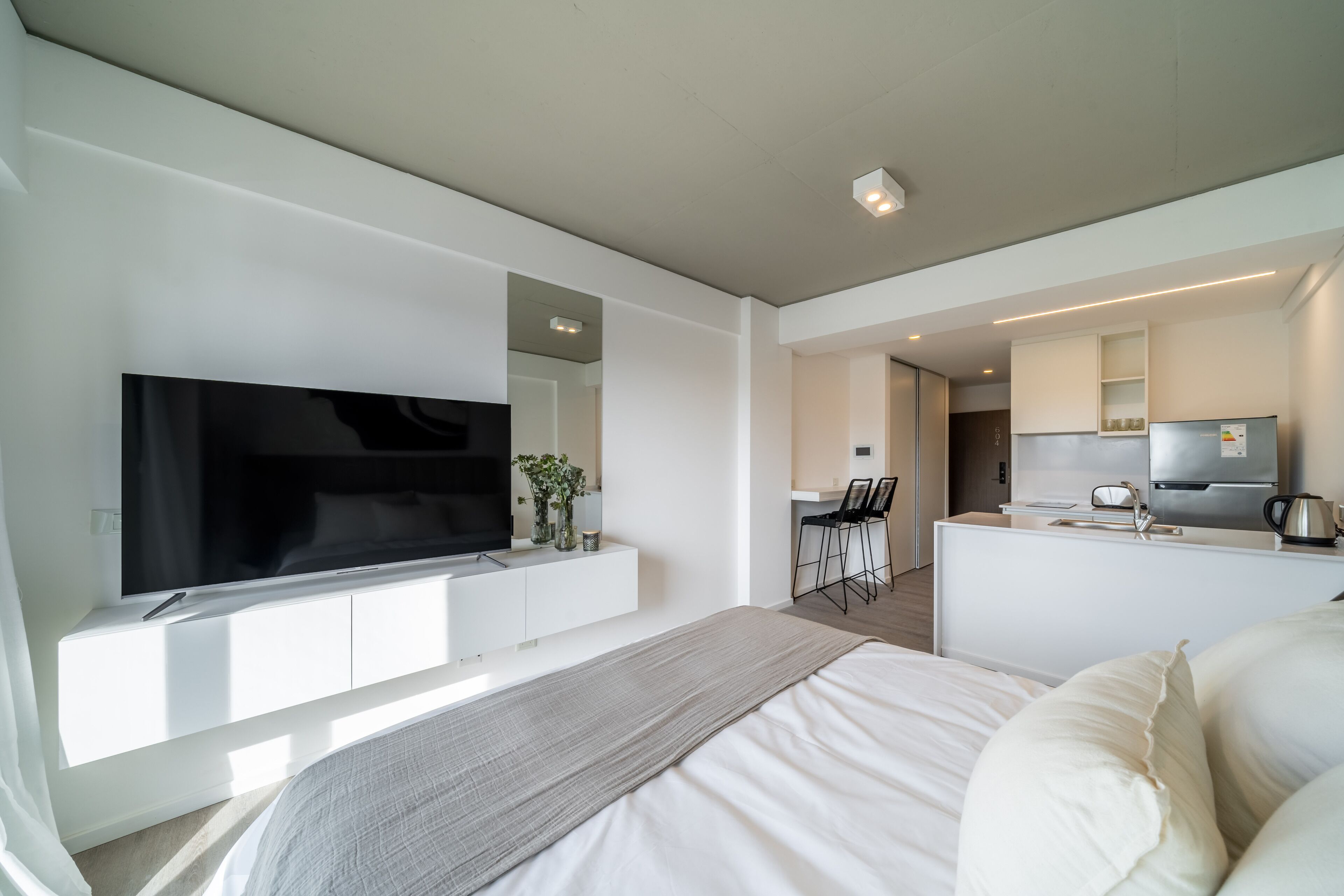 Una habitación de hotel moderna con una cama grande, un televisor de pantalla plana montado en la pared, una kitchenette y un pequeño comedor.