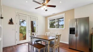 A dining area with a wooden table and chairs, a ceiling fan, a refrigerator, and a window with a view of trees. - Wandering Pines - Scenic and peaceful getaway (Pine)